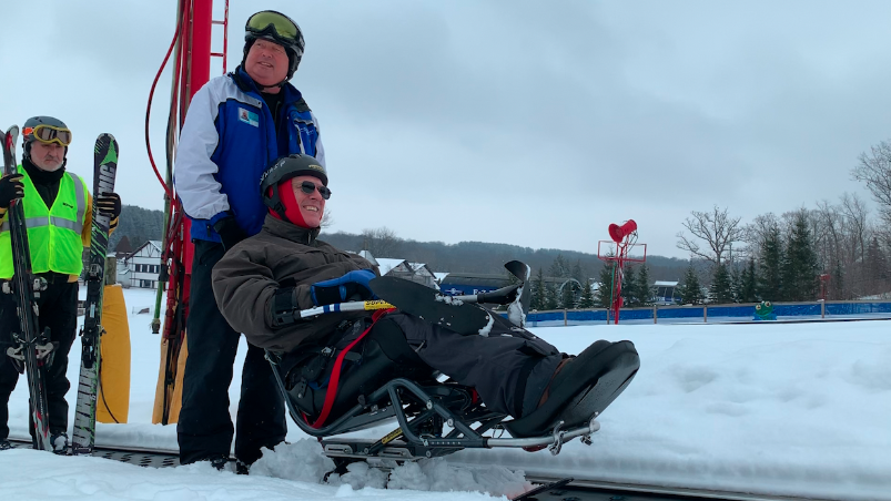 volunteer instructor riding up the magic carpet with smiling Veteran in biski with handheld outriggers